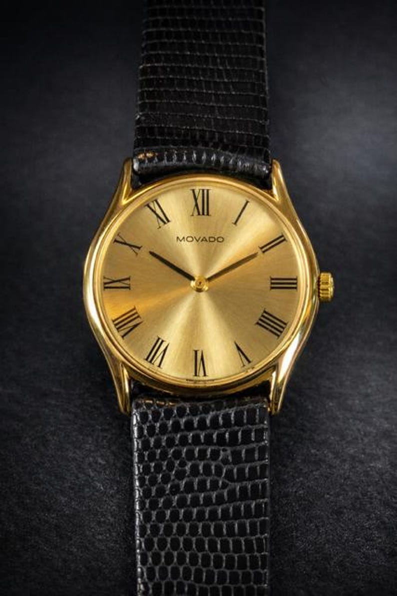 May include: Gold-toned Movado watch with a black leather band. The watch face features Roman numerals and the Movado logo. The band has a textured, reptilian pattern. The watch has a classic, elegant design.