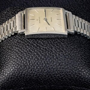 May include: A vintage silver wristwatch with a rectangular face and a silver link band. The watch face is a light cream color with black hands and markers. The watch is marked "Eterna-Matic" and "Sahida".