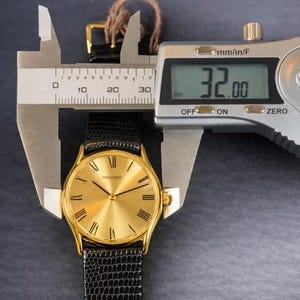 May include: A gold-toned Movado watch with a black textured band is measured with a digital caliper. The watch face has Roman numerals and a gold finish. The caliper displays a measurement of 32.00 mm.