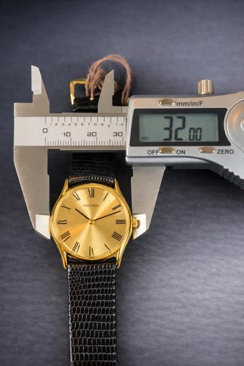 May include: A gold-toned Movado watch with a black textured band is measured with a digital caliper. The watch face has Roman numerals and a gold finish. The caliper displays a measurement of 32.00 mm.