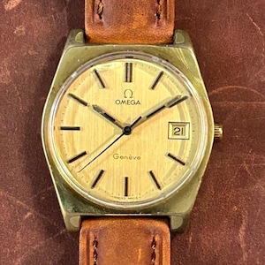 May include: A vintage Omega Genève watch with a gold-toned face and hands, featuring a date window at the 3 o'clock position. The watch has a gold-colored case and a brown leather strap. The watch face displays the Omega logo and the word "Genève".