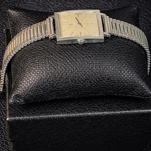 May include: A vintage wristwatch with a square, light-colored face and silver-toned band. The watch is displayed on a black leatherette pillow and box, showcasing its classic design and craftsmanship. The watch face has a small second hand.