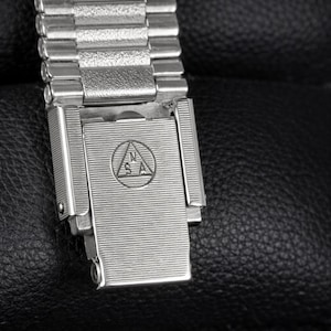 May include: Close-up of a silver-toned watch band and clasp. The band is made of small, rectangular metal links. The clasp has a textured surface with an engraved emblem. The watch band is on a black leather surface.