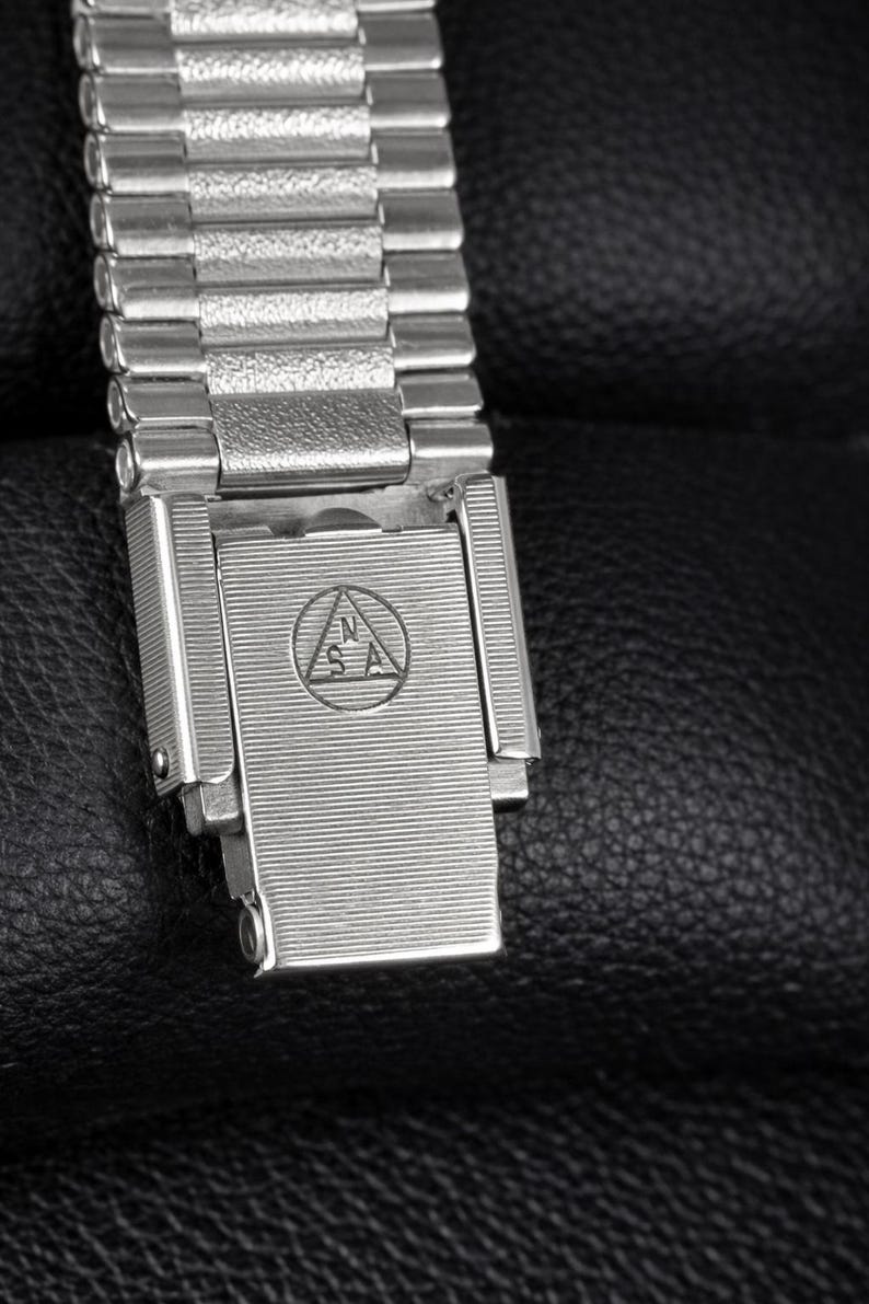 May include: Close-up of a silver-toned watch band and clasp. The band is made of small, rectangular metal links. The clasp has a textured surface with an engraved emblem. The watch band is on a black leather surface.