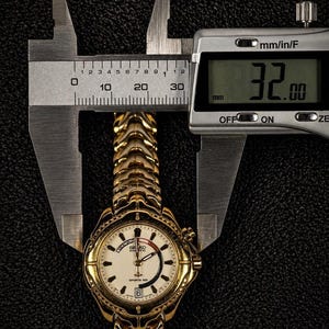 May include: A gold-toned Seiko Sports 100 wristwatch, measured at 32 mm, is displayed between the jaws of a digital caliper. The watch features a white face with black numerals and hands, and a gold-colored band. The background is a textured black surface.