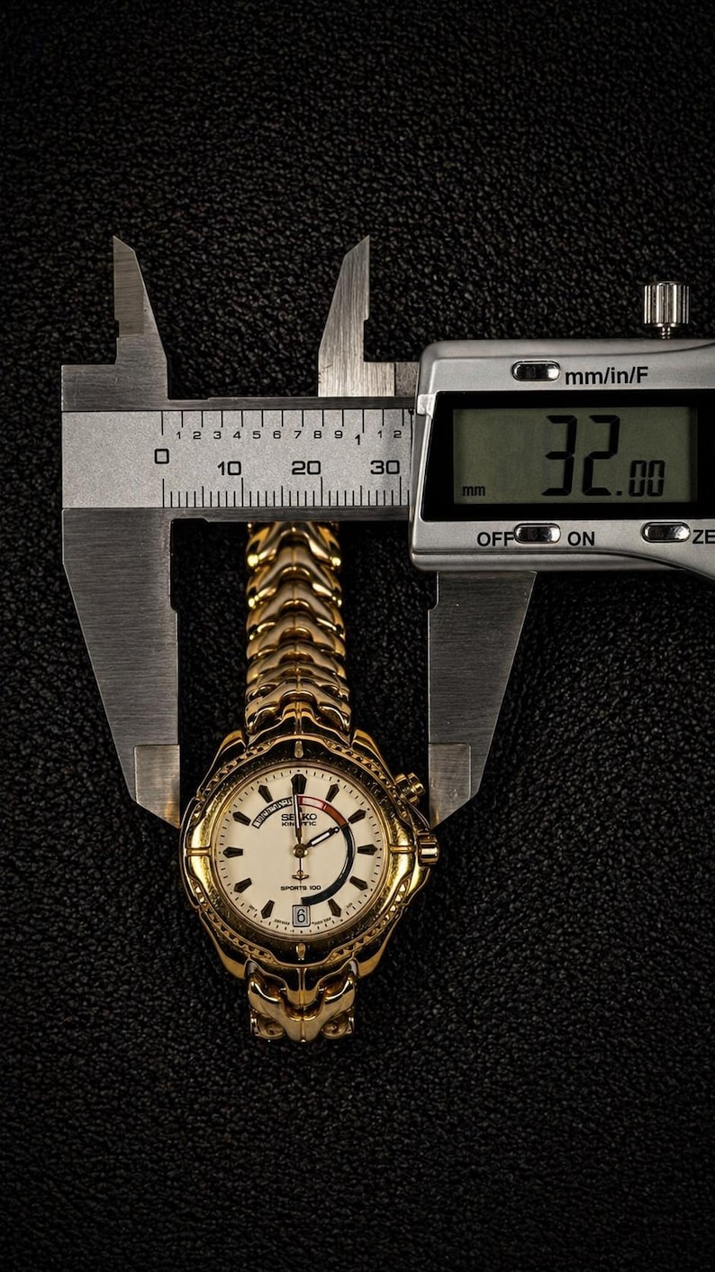 May include: A gold-toned Seiko Sports 100 wristwatch, measured at 32 mm, is displayed between the jaws of a digital caliper. The watch features a white face with black numerals and hands, and a gold-colored band. The background is a textured black surface.