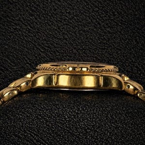 May include: A gold-toned wristwatch with a matching band, viewed from the side. The watch face is dark, and the bezel has a decorative pattern. The band links are small and rounded. The watch is set against a textured black background.