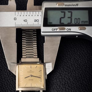 May include: A vintage Eterna Matic Sahida watch is measured with a digital caliper. The watch has a gold-colored square face and a silver band. The caliper displays a measurement of 23.00 mm.