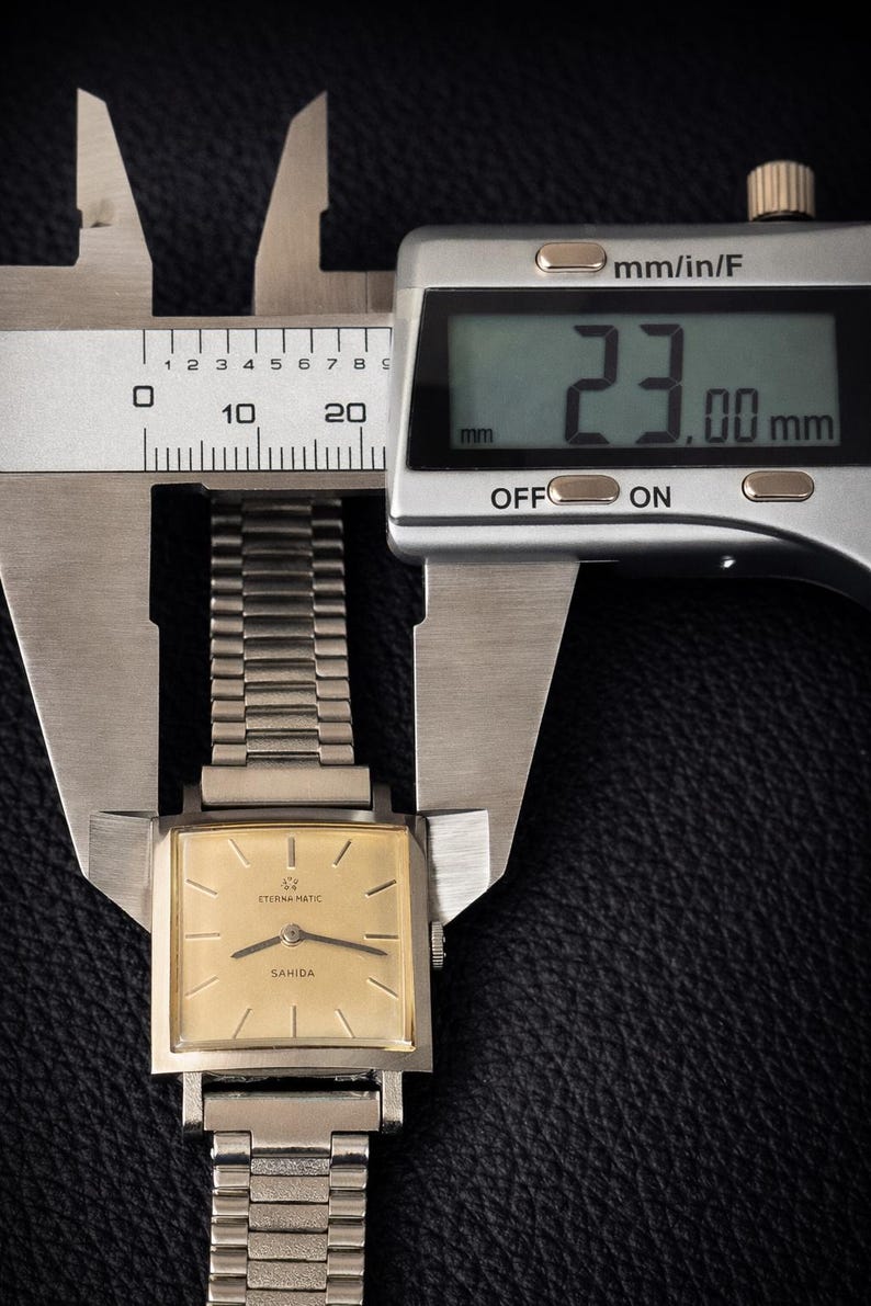 May include: A vintage Eterna Matic Sahida watch is measured with a digital caliper. The watch has a gold-colored square face and a silver band. The caliper displays a measurement of 23.00 mm.