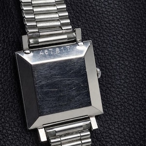 May include: A close-up of a silver wristwatch with a rectangular face and a metal band. The watch face has the number "467817" engraved on it. The watch is set against a black leather background.