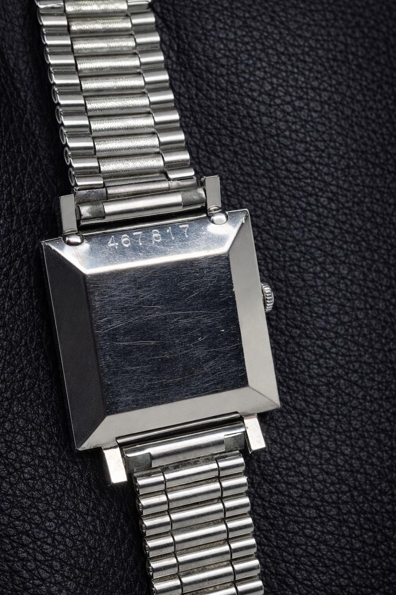 May include: A close-up of a silver wristwatch with a rectangular face and a metal band. The watch face has the number "467817" engraved on it. The watch is set against a black leather background.