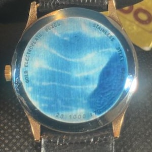 May include: Close-up of a gold-toned watch with a black leather band. The watch face has a blue design. The back of the watch is visible, with text that reads "Gold Electroplate Bezel" and "Stainless Steel Back".
