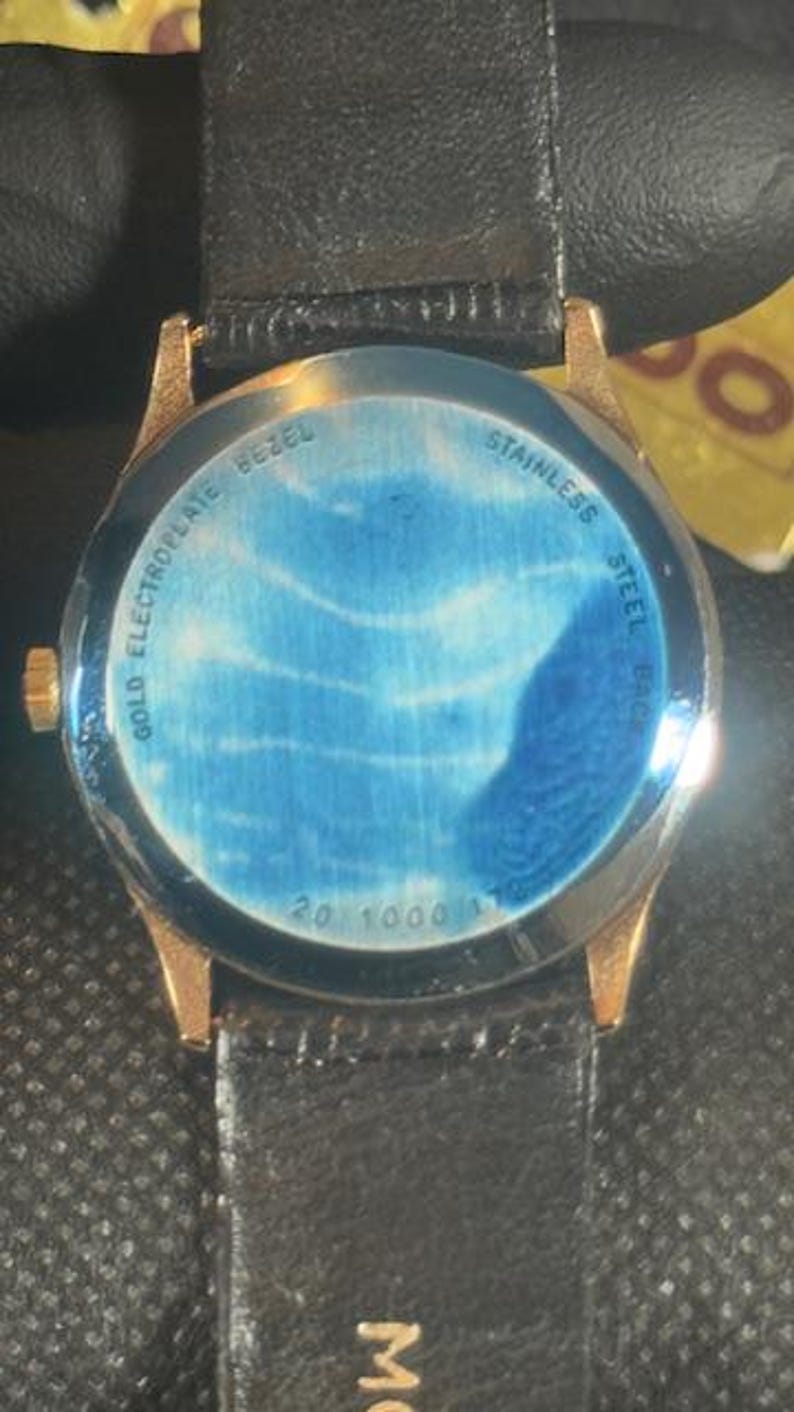 May include: Close-up of a gold-toned watch with a black leather band. The watch face has a blue design. The back of the watch is visible, with text that reads "Gold Electroplate Bezel" and "Stainless Steel Back".