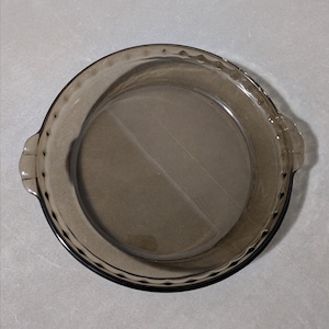 May include: A round, transparent, smoke-colored glass pie dish featuring a scalloped edge design and two integrated side handles. The dish has a smooth, circular interior base and a decorative, fluted rim that extends around the entire perimeter.