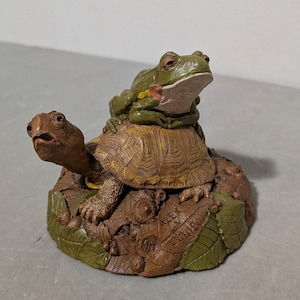 May include: A decorative figurine featuring a green frog perched on the shell of a brown turtle. The animals rest on a textured base of brown leaves and acorns, with the text "ALEX" and "LOUIE" and the number "94" engraved on the surface.