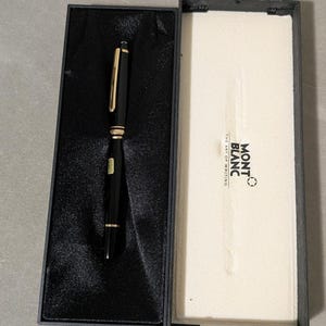 May include: A black fountain pen with gold-colored accents rests inside a black rectangular box with a black satin lining. The box lid is open, revealing a white interior panel printed with the text MONT BLANC THE ART OF WRITING.