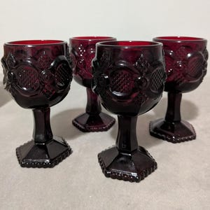 May include: A set of four deep red glass goblets featuring a raised geometric pattern on the bowl, a thick stem, and a hexagonal base. The pressed glass design includes circular and diamond-shaped textures around the exterior of each vessel.