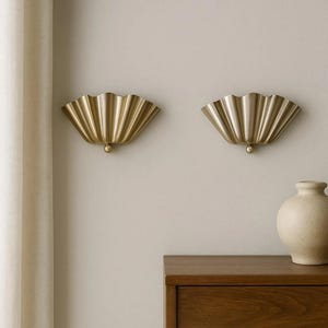May include: Two gold-coloured, fan-shaped wall sconces with a scalloped design are mounted on a light beige wall. Each sconce has a small, round detail at the bottom. A wooden cabinet and a beige vase are in the background.