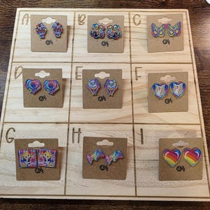 May include: A wooden display board featuring nine pairs of colourful earrings. Each pair is attached to a small cardboard tag. Designs include butterflies, hearts, and abstract shapes. The earrings are in various colours, including rainbow and pastel shades.