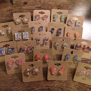May include: A collection of colorful anime-style earrings displayed on brown cardboard earring cards. The earrings feature various characters and designs, including figures, books, and scenes. The earrings are on a wooden surface.