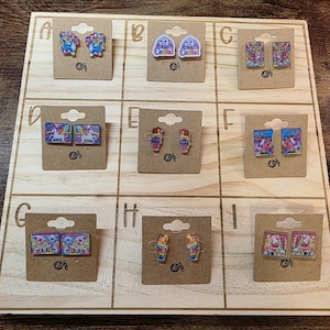 May include: A wooden display board showcasing nine pairs of novelty earrings. The earrings feature diverse designs, including unicorns, bunnies, and ice cream cones. Each pair is attached to a small, brown cardboard backing.
