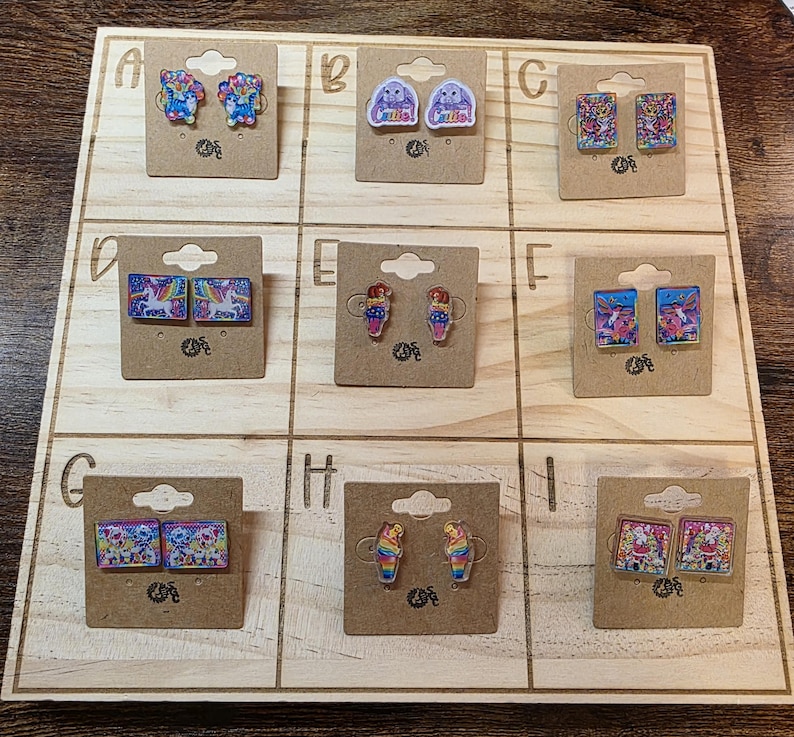 May include: A wooden display board showcasing nine pairs of novelty earrings. The earrings feature diverse designs, including unicorns, bunnies, and ice cream cones. Each pair is attached to a small, brown cardboard backing.