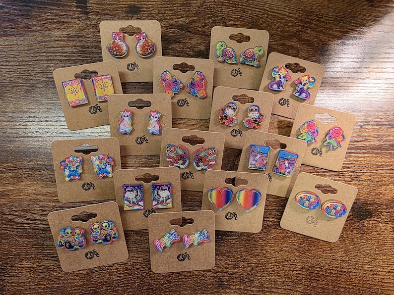 May include: A selection of colourful novelty earrings presented on brown cardboard earring cards. The earrings feature various designs, including animals, books, and abstract shapes. The earrings are made of a shiny material and are set against a wooden background.