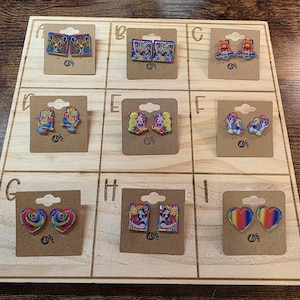 May include: A wooden display board showcasing nine pairs of earrings. The earrings display diverse designs, such as hearts, bears, and animal prints. Each pair is affixed to a small, brown card.