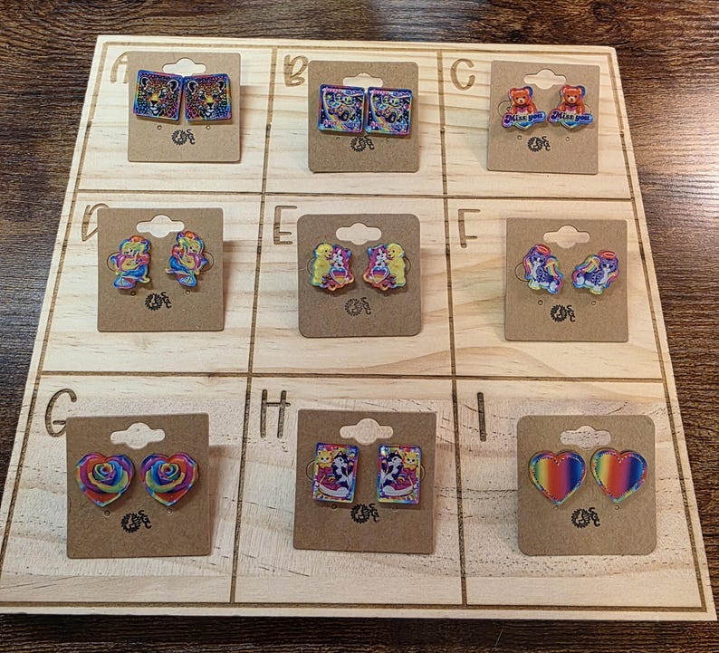 May include: A wooden display board showcasing nine pairs of earrings. The earrings display diverse designs, such as hearts, bears, and animal prints. Each pair is affixed to a small, brown card.