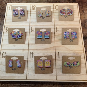May include: A wooden display board showcasing nine pairs of decorative earrings. The earrings feature diverse designs, including unicorns, fairies, cats, and turtles. Each pair is mounted on a small, brown cardboard backing.