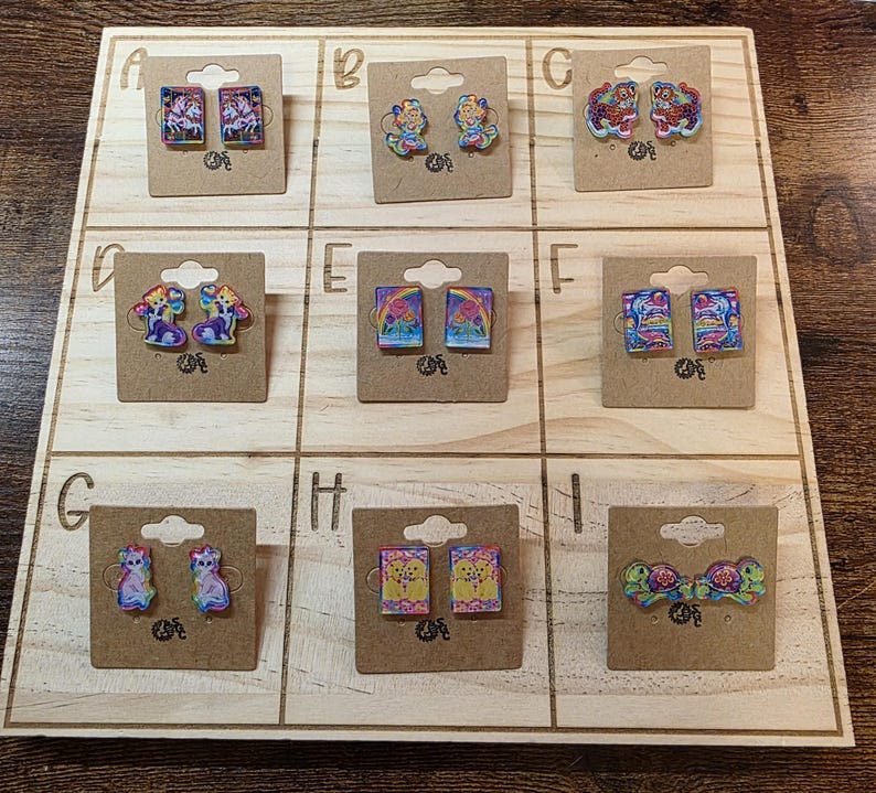 May include: A wooden display board showcasing nine pairs of decorative earrings. The earrings feature diverse designs, including unicorns, fairies, cats, and turtles. Each pair is mounted on a small, brown cardboard backing.