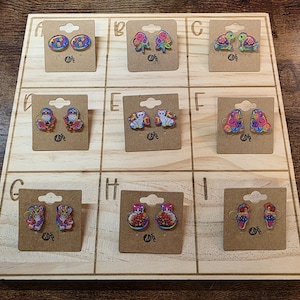 May include: A wooden display board showcasing nine pairs of decorative earrings. The earrings display diverse designs, such as flowers, turtles, owls, and abstract patterns. Each pair is mounted on a small, brown cardboard backing.