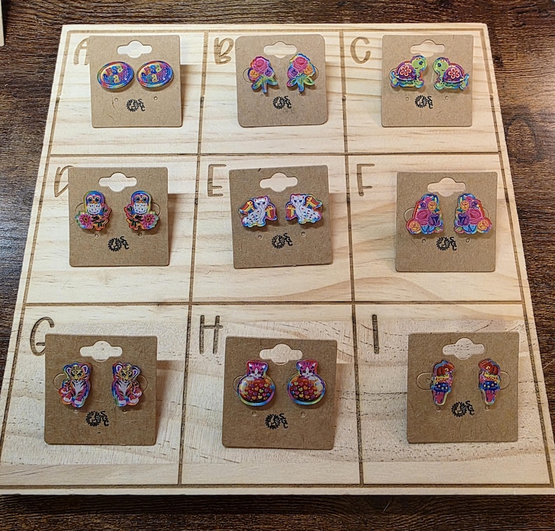 May include: A wooden display board showcasing nine pairs of decorative earrings. The earrings display diverse designs, such as flowers, turtles, owls, and abstract patterns. Each pair is mounted on a small, brown cardboard backing.
