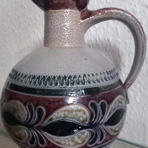 Może przedstawiać: Ceramiczny dzbanek o zaokrąglonym korpusie z jednym uchwytem. Naczynie posiada teksturowaną beżową górną część oraz ciemnośliwkową dolną część ozdobioną powtarzającym się czarno-beżowym wzorem liści oraz poziomymi pasami.
