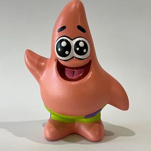 May include: A pink starfish figurine with large, expressive eyes and a wide, smiling mouth. The character is wearing green and purple shorts. The figurine is a cartoon character, likely from a popular animated series.