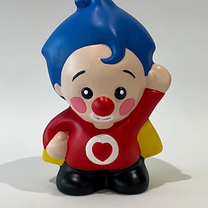 May include: A figurine of a cartoon character with blue hair, a red shirt with a white heart, and a yellow cape. The character has a red nose, rosy cheeks, and black pants. The figurine is standing with one arm raised.