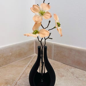 Könnte beinhalten: Eine schwarze, tropfenförmige Vase mit pfirsichfarbenen Kunstblumen mit grünen Zentren. Die Vase hat ein modernes, minimalistisches Design. Die Blumen haben zarte Blütenblätter und sind so angeordnet, dass sie eine natürliche Ästhetik erzeugen. Die Vase steht auf einer gefliesten Oberfläche.