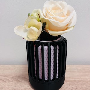 Puede incluir: Un jarrón decorativo negro con listones verticales, que contiene un interior rosa claro y un ramo de flores. Las flores incluyen una gran rosa blanca cremosa y flores amarillas pálidas más pequeñas, creando un arreglo suave y elegante.