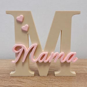 May include: A decorative wooden letter "M" with the word "Mama" in pink script. Two pink hearts are attached to the upper left side of the letter. The letter is beige.