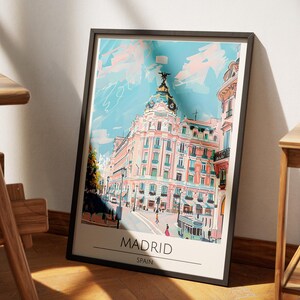 May include: A framed art print featuring a colorful illustration of a building in Madrid, Spain. The artwork showcases a detailed architectural rendering with a light blue sky and pink accents. The word "MADRID" is printed at the bottom of the print.