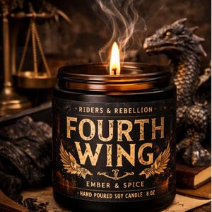 Op de afbeelding: Een brandende, handgegoten sojakaars in een zwarte pot met de tekst "FOURTH WING" en "EMBER & SPICE". De kaars is 227g en heeft een gouden rand. De kaars rookt en een drakenbeeld staat op de achtergrond.
