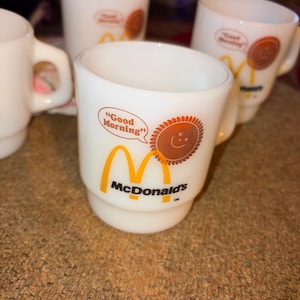 May include: A set of white milk glass mugs featuring the McDonald's logo in yellow and black text. Each mug displays a brown sun graphic with a smiling face and a speech bubble containing the text Good Morning in red letters on the front side.