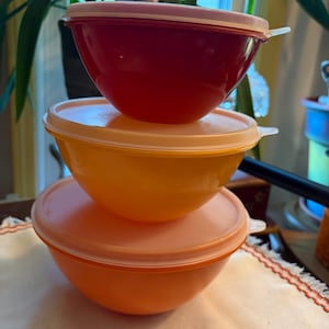 Puede incluir: Una pila de tres cuencos redondos de plástico con tapas a juego. El cuenco superior es rojo, el del medio es amarillo y el inferior es naranja. Cada cuenco presenta un diseño liso y curvado con una pequeña pestaña en la tapa para abrir.