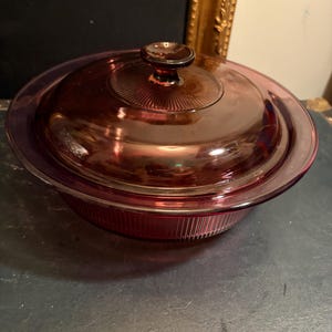Puede incluir: Una fuente de horno redonda de cristal de color morado oscuro con tapa a juego. El recipiente presenta detalles acanalados verticales en los laterales exteriores, mientras que la tapa tiene un pomo central rodeado por un patrón circular de líneas radiantes.