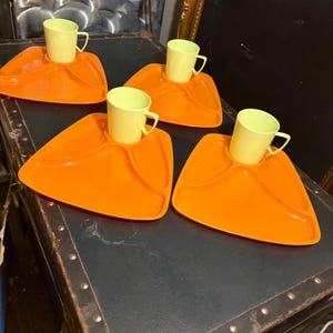 May include: A set of four bright orange triangular snack trays, each featuring a dedicated circular indentation for a matching pale yellow cup. The trays have rounded corners and a smooth finish, with each cup positioned in the center of the tray.