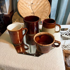 May include: A collection of brown ceramic mugs and a small jar on a mirrored surface. The mugs vary in size and design, with some having a lighter-colored rim. A woven hat is in the background.
