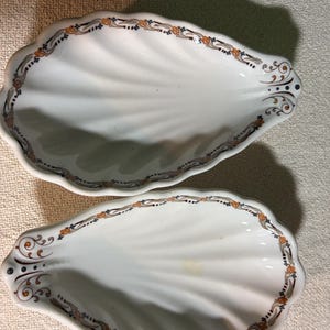 May include: Two white, shell-shaped ceramic dishes with a decorative border of orange, brown, and blue accents. The dishes have a scalloped edge and a detailed, swirling pattern on the interior.