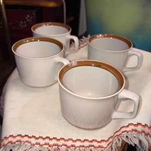 May include: A set of four off-white ceramic mugs, each featuring a smooth, rounded body and a simple handle. The top rim of every mug is accented with a distinct, solid brown band, providing a clean contrast against the light-colored base.