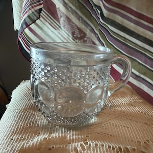 May include: Clear glass teacup with a handle. The cup features a textured pattern of small circles and larger floral designs. It rests on a white lace tablecloth, with a striped fabric in the background.