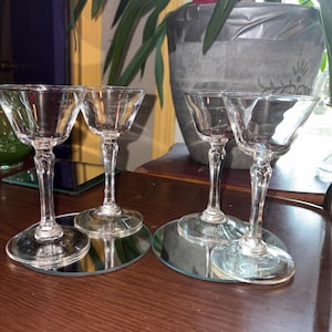 May include: Four clear glass stemmed cordial glasses with wide, flared bowls and faceted stems. Each glass features a circular base and a smooth, transparent finish. The set is arranged in two pairs, with each pair resting on a circular mirror coaster.